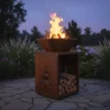 Brasero acier corten avec feu allumé et espace de rangement bois intégré dans jardin ambiance crépusculaire